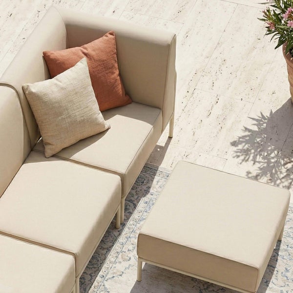 Modulare Gartenlounge mit Sofa und Hocker, Polsterung in hellem Stoff, zwei Dekokissen, auf einer Terrasse mit Natursteinfliesen und Außenteppich.