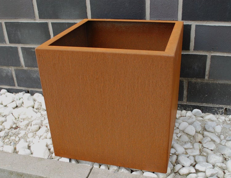 Quadratischer Pflanzkübel aus Cortenstahl in Rostoptik auf weißen Kieselsteinen vor einer dunklen Mauer.