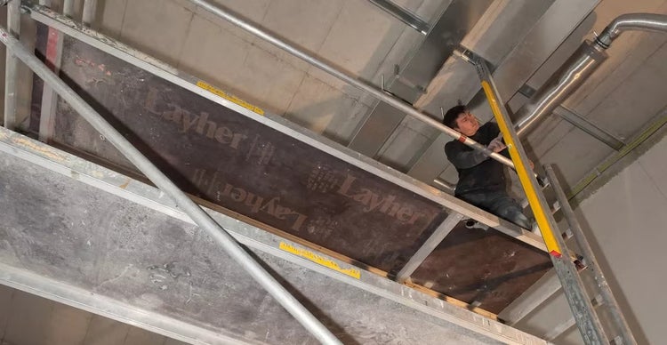 Handwerker arbeitet auf einem Layher Fassadengerüst an der Decke eines Rohbaus mit Lüftungsrohren.