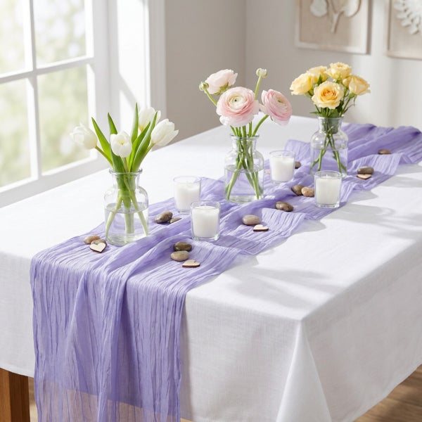 Tischdekoration mit einem fliederfarbenen Tischläufer aus Stoff, Glasvasen mit Blumen, Kerzengläsern und dekorativen Steinen auf einem weißen Tisch.