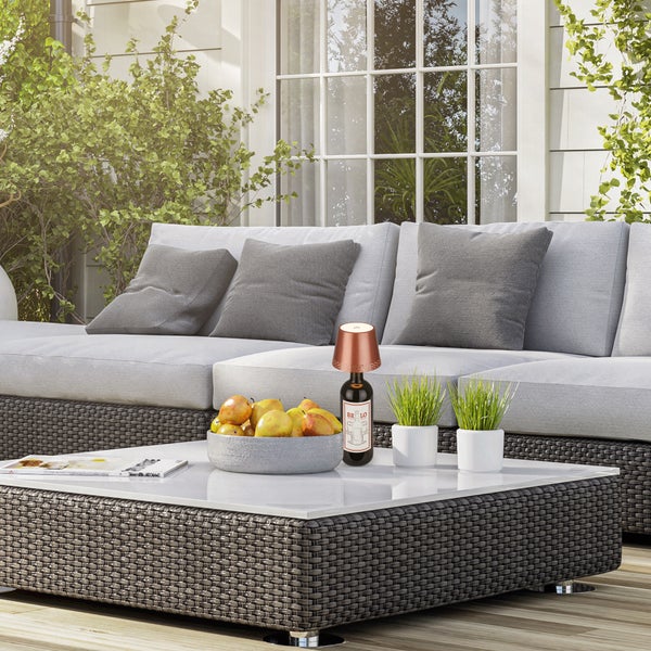 Moderne Gartenlounge aus grauem Kunststoffgeflecht mit Sofa und Tisch auf einer hellen Terrasse, dekoriert mit Kissen, Obstschale und Flaschenleuchte.