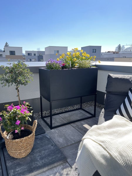 Helle, organisierte Terrasse mit einem modernen schwarzen Hochbeet aus Metall, bunten Blumen, einem Loungesessel und einem Weidenkorb.