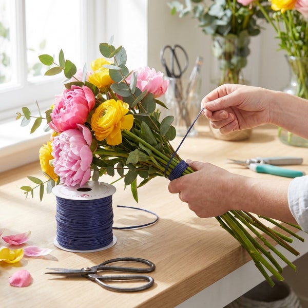 Eine Person bindet einen Blumenstrauß aus Pfingstrosen und Ranunkeln mit einer Schnur von einer Spule auf einem Holztisch.