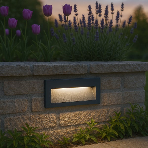 Rechteckige Wandeinbauleuchte für den Außenbereich in einer Natursteinmauer mit warmem Lichtstrahl nach unten auf Pflanzen.