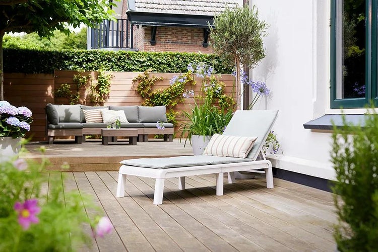 Helle Terrasse mit Holzdielen, weißer Sonnenliege mit grauen Kissen und Holz-Lounge-Möbeln im Hintergrund.