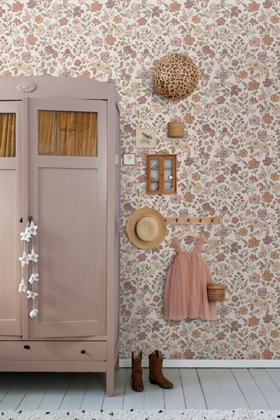Hell gestaltetes Kinderzimmer mit floral gemusterter Vliestapete, einem rosa Holzschrank im Landhausstil und verschiedenen Naturdekorationen.