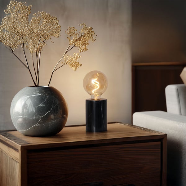 Moderne Tischleuchte mit schwarzem Marmorfuß und leuchtender Filamentlampe neben einer grauen Marmorvase auf einem Holztisch.