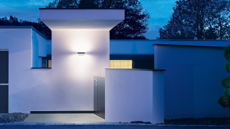 Moderne Außenwandleuchte an einer weißen Hausfassade bei Dämmerung, die den Eingangsbereich hell ausleuchtet.