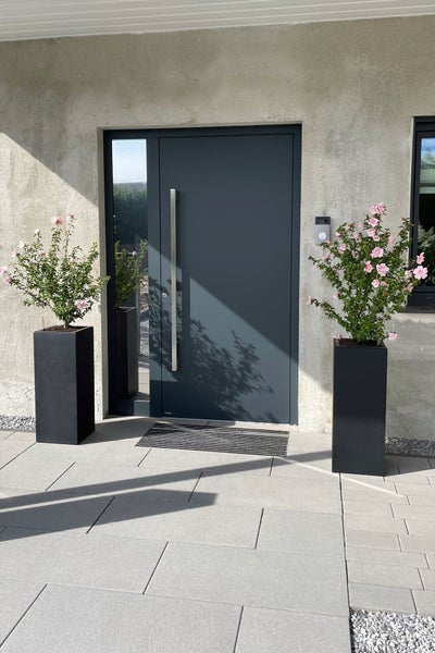 Moderner Hauseingang mit anthrazitfarbener Haustür, Seitenteil aus Glas, Steinplatten und hohen schwarzen Pflanzkübeln mit Blumen.