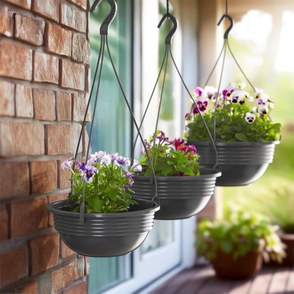 Drei hängende Pflanzkübel mit blühenden Blumen, befestigt an einer Ziegelmauer neben einem Fenster.