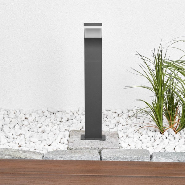 Moderne LED-Wegeleuchte aus Metall in Anthrazit, montiert auf einem Steinsockel in einer Gartenanlage mit Zierkies und Pflanzen.