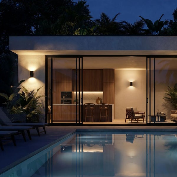 Moderne Villa bei Nacht mit beleuchtetem Pool, Wandleuchten an der Fassade und Blick durch Glasfronten in eine Küche mit Holzelementen.
