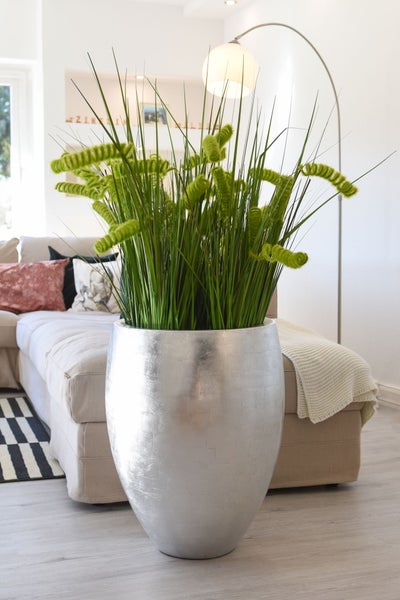 Große silberne Bodenvase mit künstlichem Gras in einem hellen Wohnzimmer mit beigem Sofa und Stehlampe.