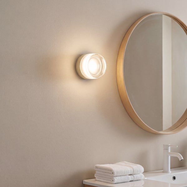 Moderne Wandleuchte in einem Badezimmer neben einem runden Spiegel mit Holzrahmen und einem Waschbecken mit Armatur und Handtüchern.