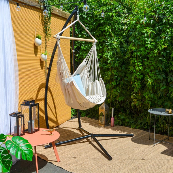 Hängesessel aus hellem Stoff mit schwarzem Metallgestell in einem gemütlichen Garten mit Teppich, Beistelltischen und Hecke.