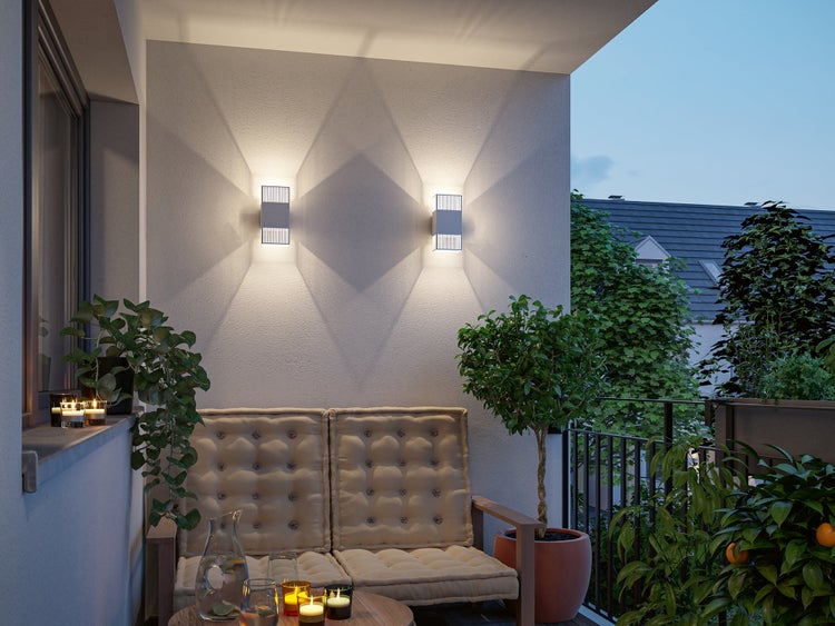 Moderner Balkon mit zwei rechteckigen Wandleuchten, einer Holzbank mit beigen Kissen, Topfpflanzen und Kerzen in der Abenddämmerung.