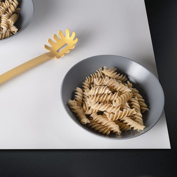 Fusilli-Nudeln in einer Keramikschüssel mit einem Nudellöffel aus Kunststoff auf einer strukturierten Unterlage.