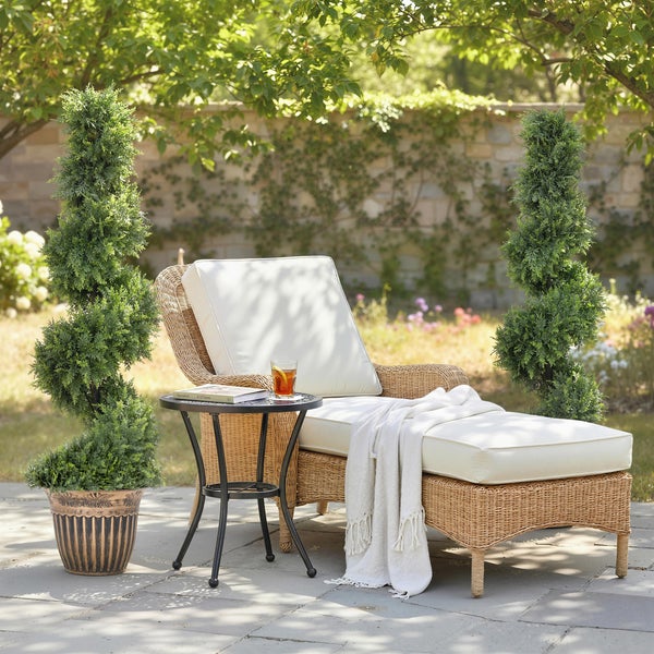 Gartenliege aus Rattan mit weicher Polsterauflage, dazu ein kleiner Beistelltisch und zwei spiralförmig geschnittene Kunstbäume in Pflanzgefäßen.