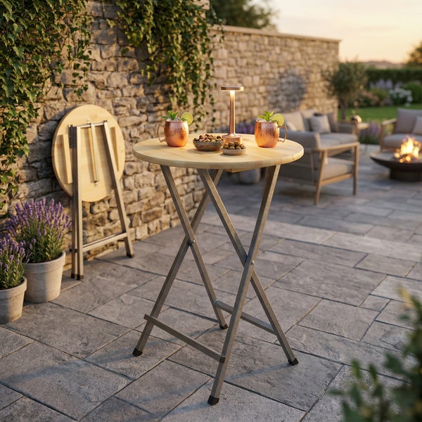 Klappbarer Stehtisch mit runder Holzplatte und Metallgestell auf einer hellen Terrasse mit Steinmauer und Gartenmöbeln.