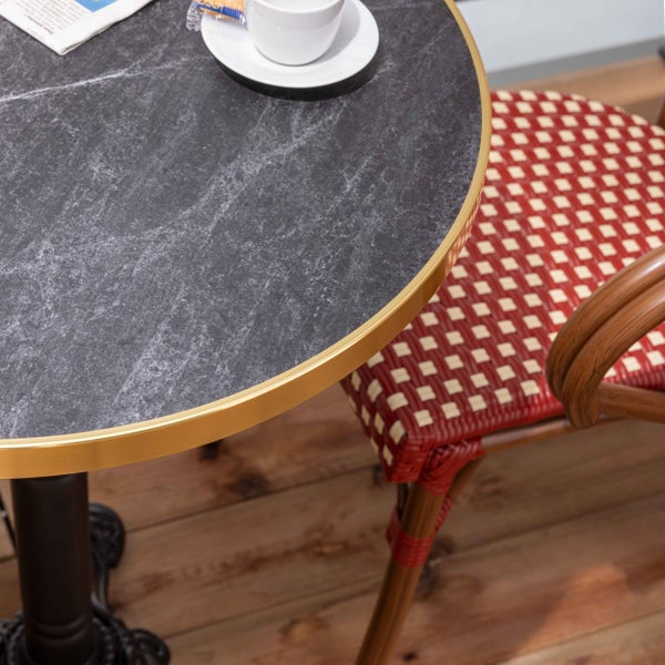 Runder Bistrotisch mit Tischplatte in dunkelgrauer Marmoroptik und goldfarbenem Rand neben einem rot-creme geflochtenen Stuhl.