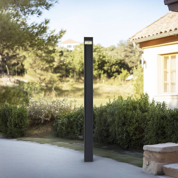 Moderne anthrazitfarbene Pollerleuchte mit gerippter Oberfläche in einem hellen Garten an einem Weg.
