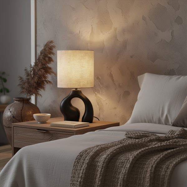 Moderne Tischleuchte mit schwarzem skulpturalem Fuß und Stoffschirm auf hölzernem Nachtschrank in einem hellen Schlafzimmer mit strukturierter Wand.