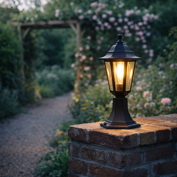 Schwarze Garten-Sockelleuchte mit warmem Licht auf einer Backsteinmauer, Garten mit Pergola und Blumen im Hintergrund.