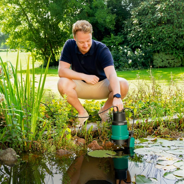 Ein Mann hockt an einem hellen Gartenteich mit Seerosen und hält eine grün-schwarze VONROC Tauchpumpe am Tragegriff über das Wasser.