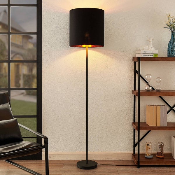 Moderne Stehlampe mit schwarzem Stoffschirm und goldenem Innenfutter in einem hellen Wohnzimmer neben einem Holzregal und Sessel.