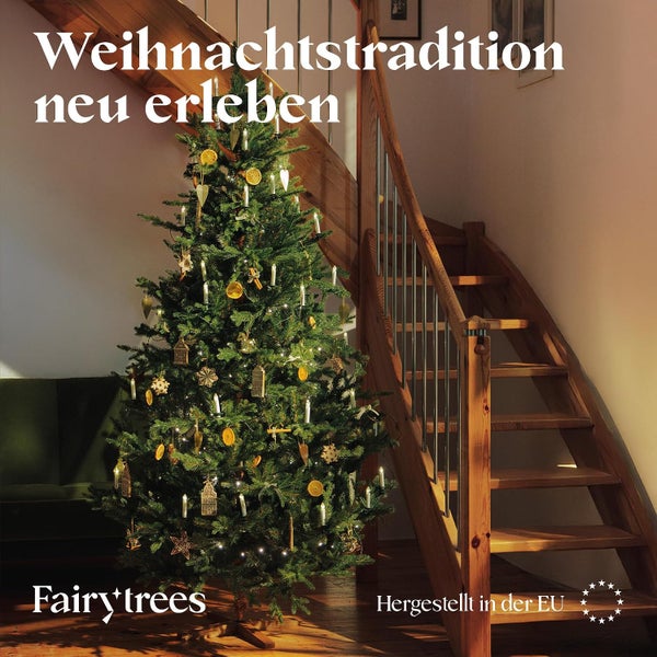 Ein geschmückter künstlicher Weihnachtsbaum von Fairy Trees steht in einem Wohnraum neben einer Holztreppe mit Lichtern und Dekorationen.