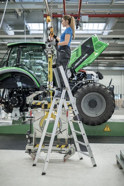 Frau auf einer Aluminium-Stufenleiter bei der Arbeit an einem Traktor in einer Werkshalle