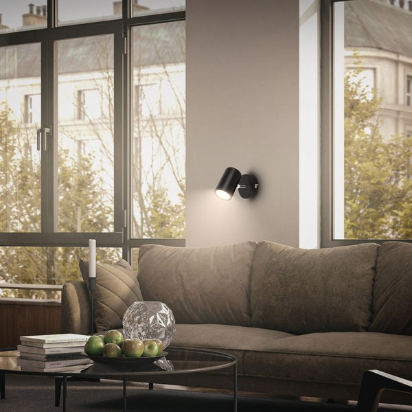 Modernes Wohnzimmer mit schwarzer Wandleuchte über einem Sofa. Helle Atmosphäre mit großen Fenstern und Glastisch.