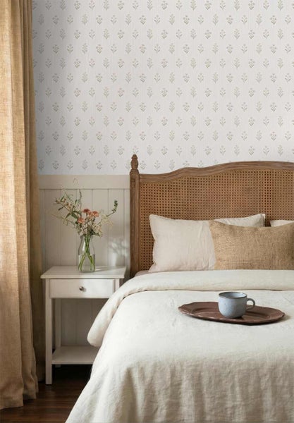 Helles Schlafzimmer mit floraler Mustertapete über Holzvertäfelung, Bett mit Geflecht-Kopfteil, Leinenbettwäsche und Nachttisch mit Blumenstrauß.