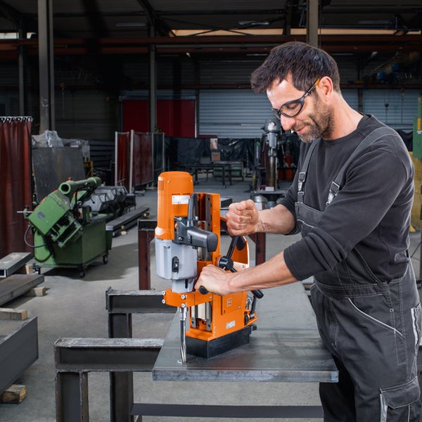 Handwerker bedient eine Magnet-Kernbohrmaschine auf einer Stahlplatte in einer hellen Werkstatt.