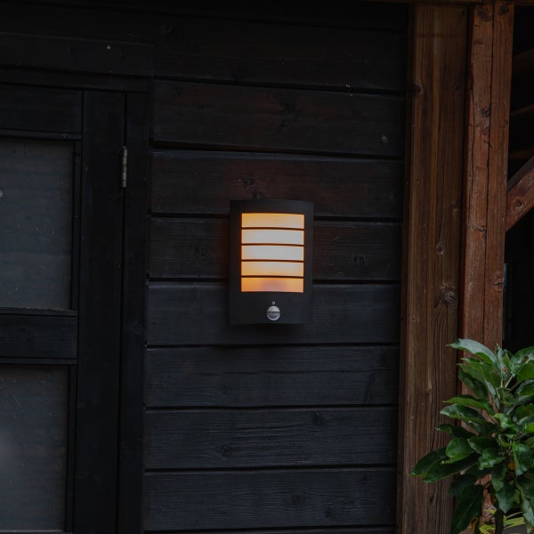 Moderne Außenwandleuchte mit Bewegungsmelder an einer dunklen Holzwand, warmes Licht strahlt durch horizontale Lamellen.