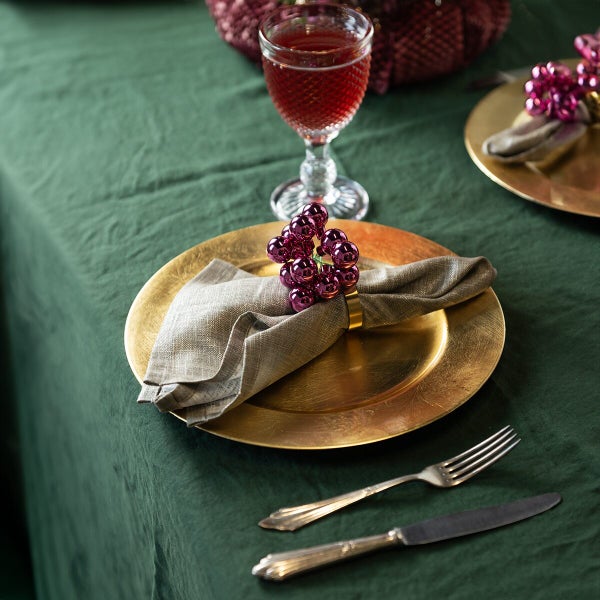 Festlich gedeckter Tisch mit goldenem Platzteller, Stoffserviette mit glänzendem Beeren Serviettenring, Weinglas und Besteck auf grüner Tischdecke.