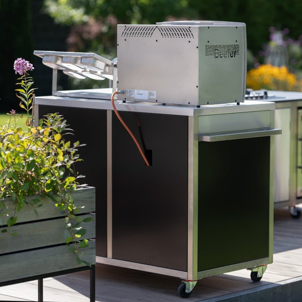 Mobiles Außenküchenmodul aus Edelstahl mit schwarzer Front und Beefer Logo Grill auf einer Holzterrasse in einem hellen Garten.