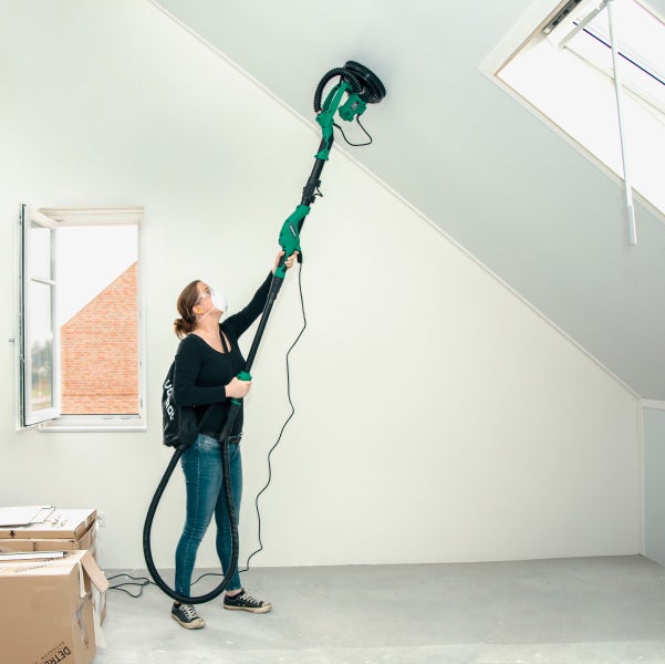 Eine Person verwendet einen Langhalsschleifer mit integriertem Staubsaugeranschluss, um eine Decke in einem ausgebauten Dachgeschoss zu schleifen.