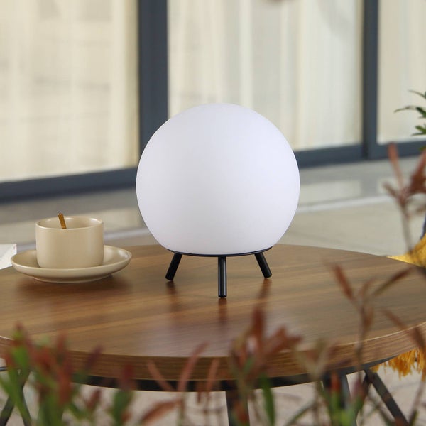 Kugelförmige Tischleuchte mit weißem Schirm und schwarzem Stativ auf einem Holztisch neben einer Tasse.