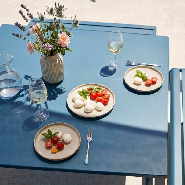 Gedeckter blauer Gartentisch aus Metall mit Tellern, Gläsern und einer Blumenvase im hellen Sonnenlicht.