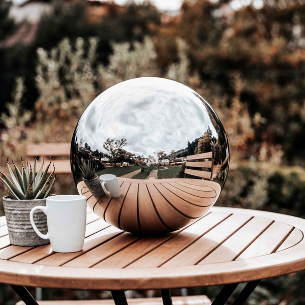 Spiegelnde Gartenkugel aus Edelstahl auf einem runden Holztisch im Freien neben einer Tasse und einer Topfpflanze.
