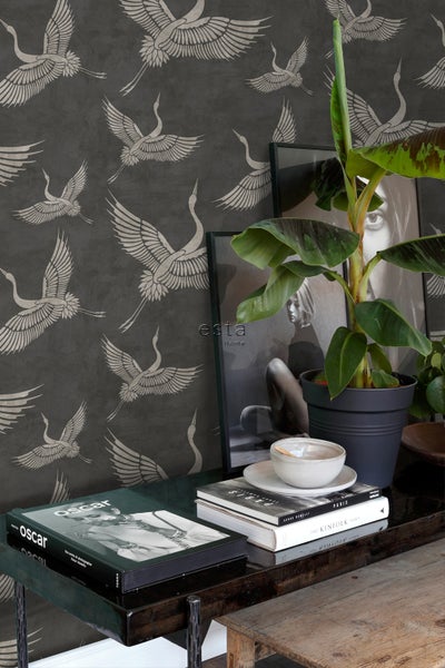 Dekoratives Zimmer mit Tapete mit Kranichmuster, Zimmerpflanze und gerahmten Bildern auf dem Tisch.