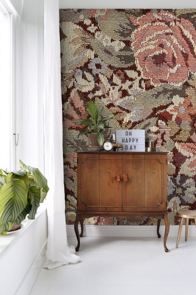 Fototapete mit großformatigem, gewebtem Blumenmuster im Tapisserie-Stil hinter einer Holzkommode in einem hellen Wohnraum.