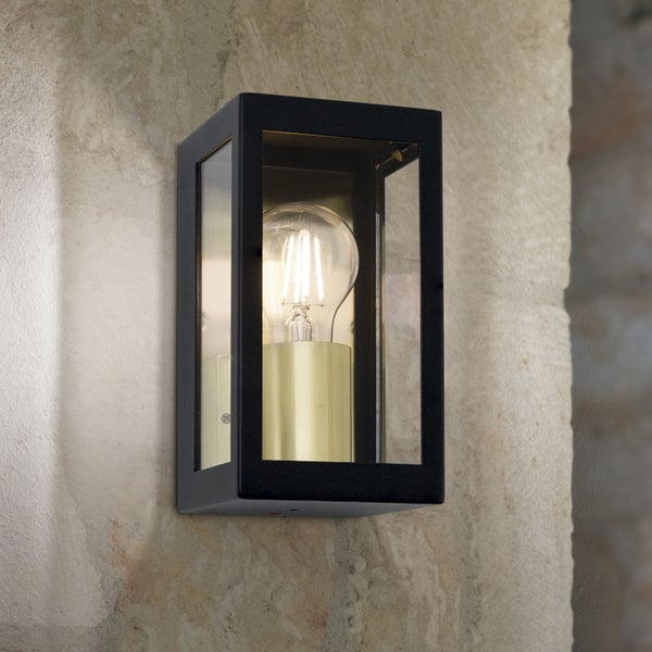 Schwarze rechteckige Außenwandleuchte mit Glaseinsätzen und goldener Fassung, montiert an einer Steinwand mit eingeschaltetem Leuchtmittel.