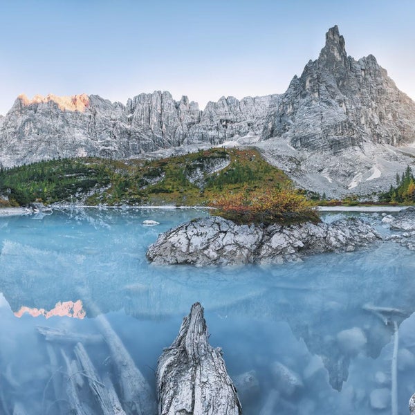 Bergsee mit Baumstämmen und Felsen im Vordergrund, Bergmassiv im Hintergrund