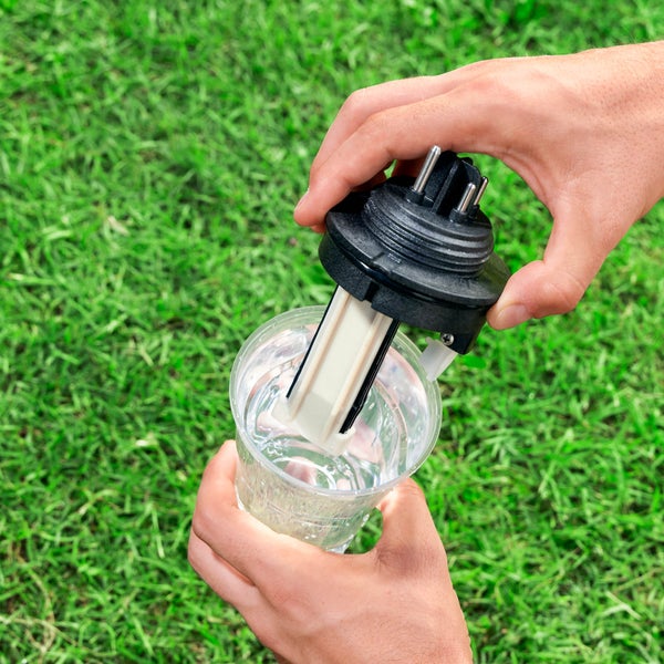 Eine Person testet ein Bewässerungssystem mit einem Messbecher und einem Feuchtigkeitssensor vor einem Rasenhintergrund.