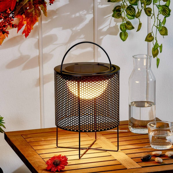 Solar-Tischleuchte aus schwarzem Lochblech mit Tragebügel auf einem Holztisch in einer hellen, dekorierten Außenszene.