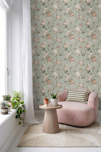 Helles Zimmer mit geblümter Tapete mit Rosenmuster, einem rosa Sessel mit gestreiftem Kissen, einem Beistelltisch aus Holz und Zimmerpflanzen.