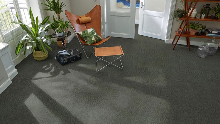 Strukturierte Teppichfliesen in einem hellen Wohnzimmer mit Ledersessel, Zimmerpflanzen und Holzregal.