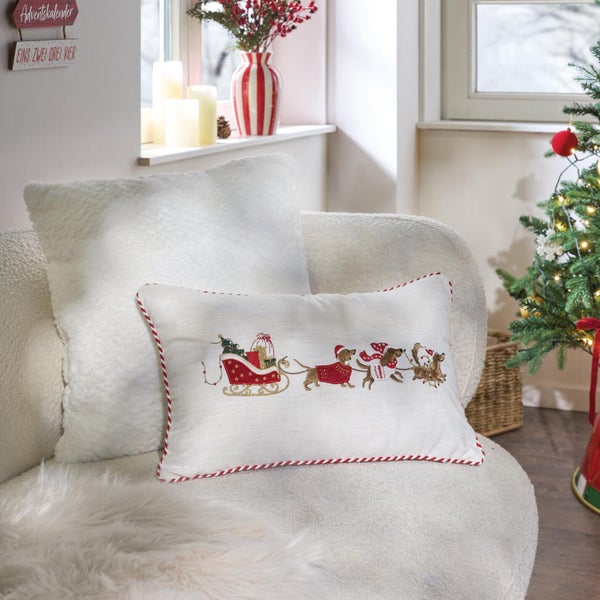 Weihnachtskissen mit Dackelmotiv und Schlittenstickerei auf hellem Untergrund als festliche Dekoration auf einem Sessel vor Weihnachtsbaum.